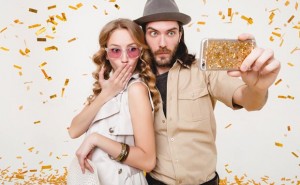 young stylish hipster couple in love making self photo, celebrating disco party, having fun, amazed, exited, funny face, golden confetti, holding phone, trendy apparel, cruise style, white background