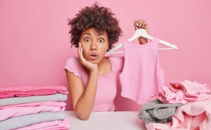 Shocked surprised woman with curly hair holds t shirt on hanger folds laundry at home does household duties cleans out closet sits at table isolated on pink background. Wardrobe analysis spring update