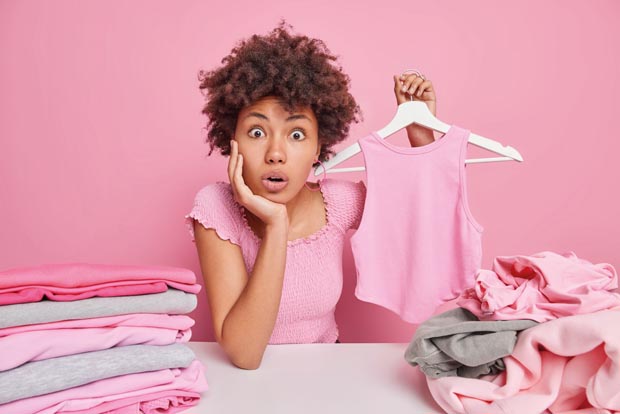 Shocked surprised woman with curly hair holds t shirt on hanger folds laundry at home does household duties cleans out closet sits at table isolated on pink background. Wardrobe analysis spring update