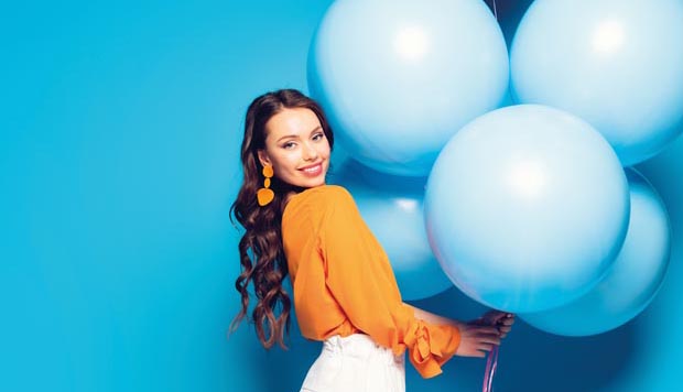 full length view of happy stylish woman standing on one leg while holding big festive balloons on blue background